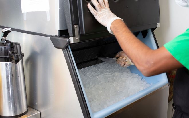 A person wearing a glove and green shirt uses a scoop to collect ice from an open commercial ice machine.
