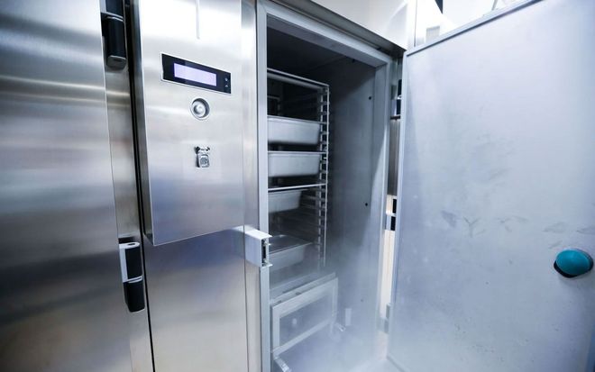 A stainless steel industrial blast chiller with an open door, revealing trays and a visible mist of cold air.