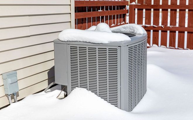 A gray outdoor HVAC unit sits next to a house wall and wooden fence, covered in a light layer of snow.