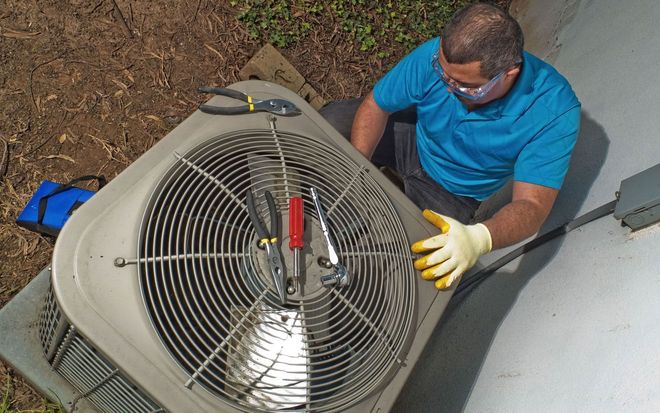 A technician wearing safety glasses and yellow gloves performs maintenance on an outdoor air conditioning unit.
