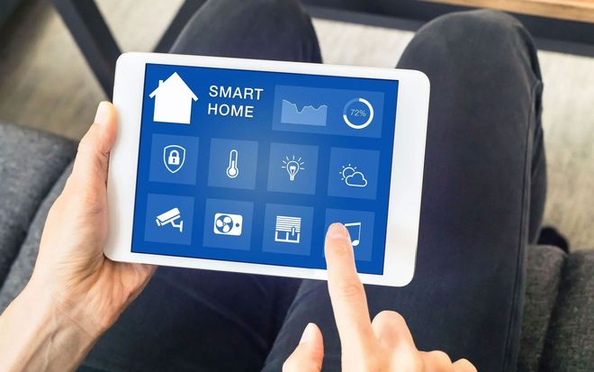 Hands holding a tablet displaying a smart home control interface with icons for security, lighting, and climate settings.