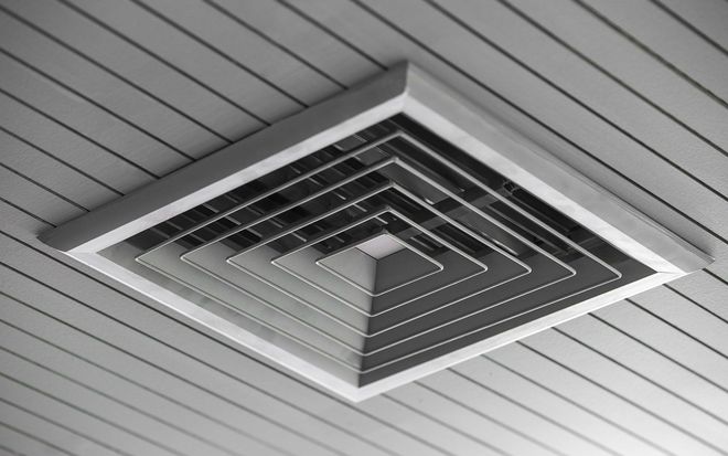 A square, multi-tiered ceiling vent with nested louvers mounted on a white, slatted panel ceiling.