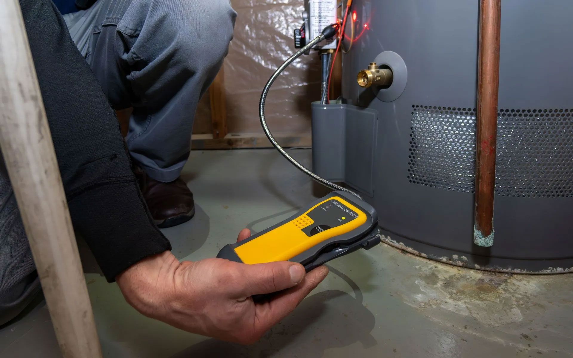A person uses a handheld yellow gas detector to check for leaks around a residential water heater in a basement.
