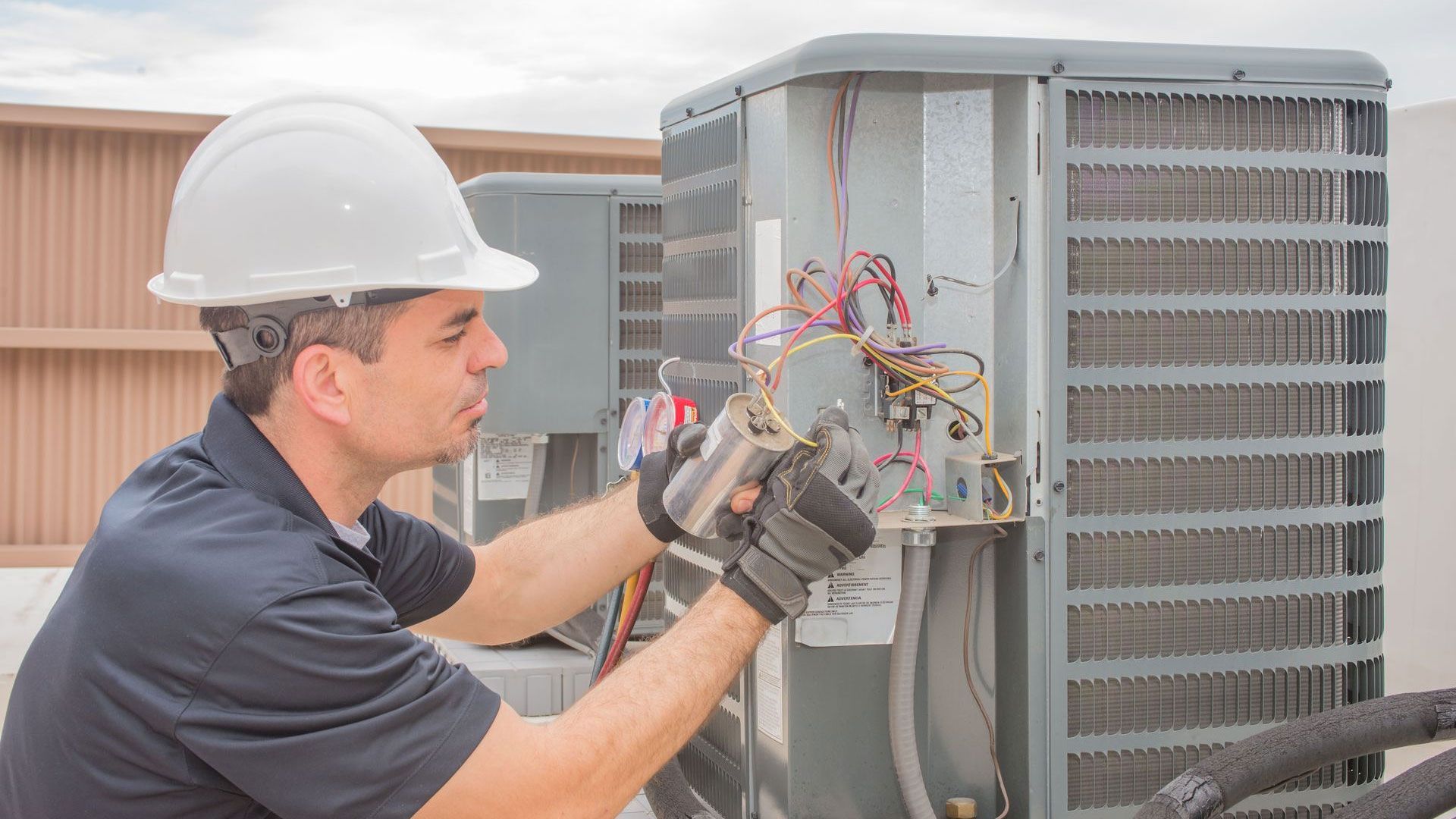 Technician wearing a hard hat and gloves repairs the electrical wiring of an outdoor air conditioning unit.
