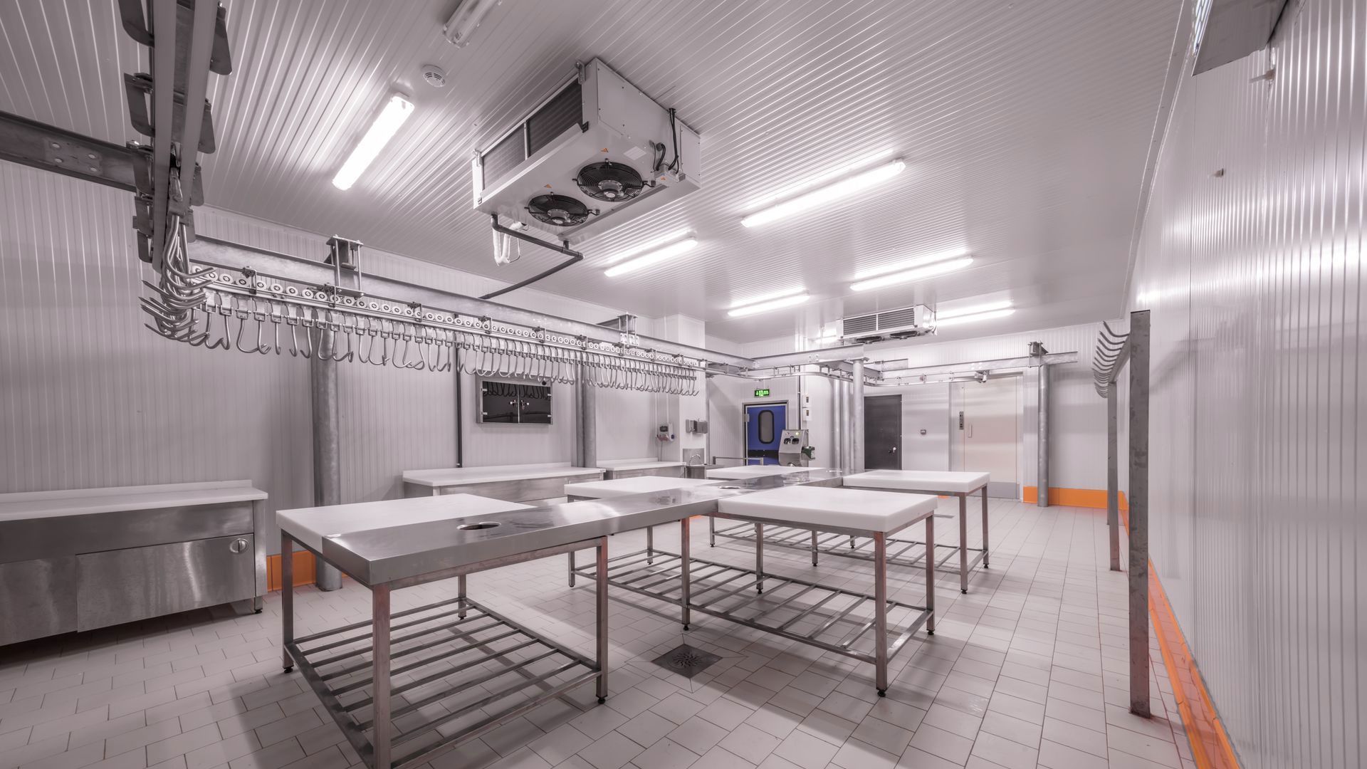 A sterile, brightly lit commercial meat processing room featuring stainless steel tables and a ceiling-mounted rail system.