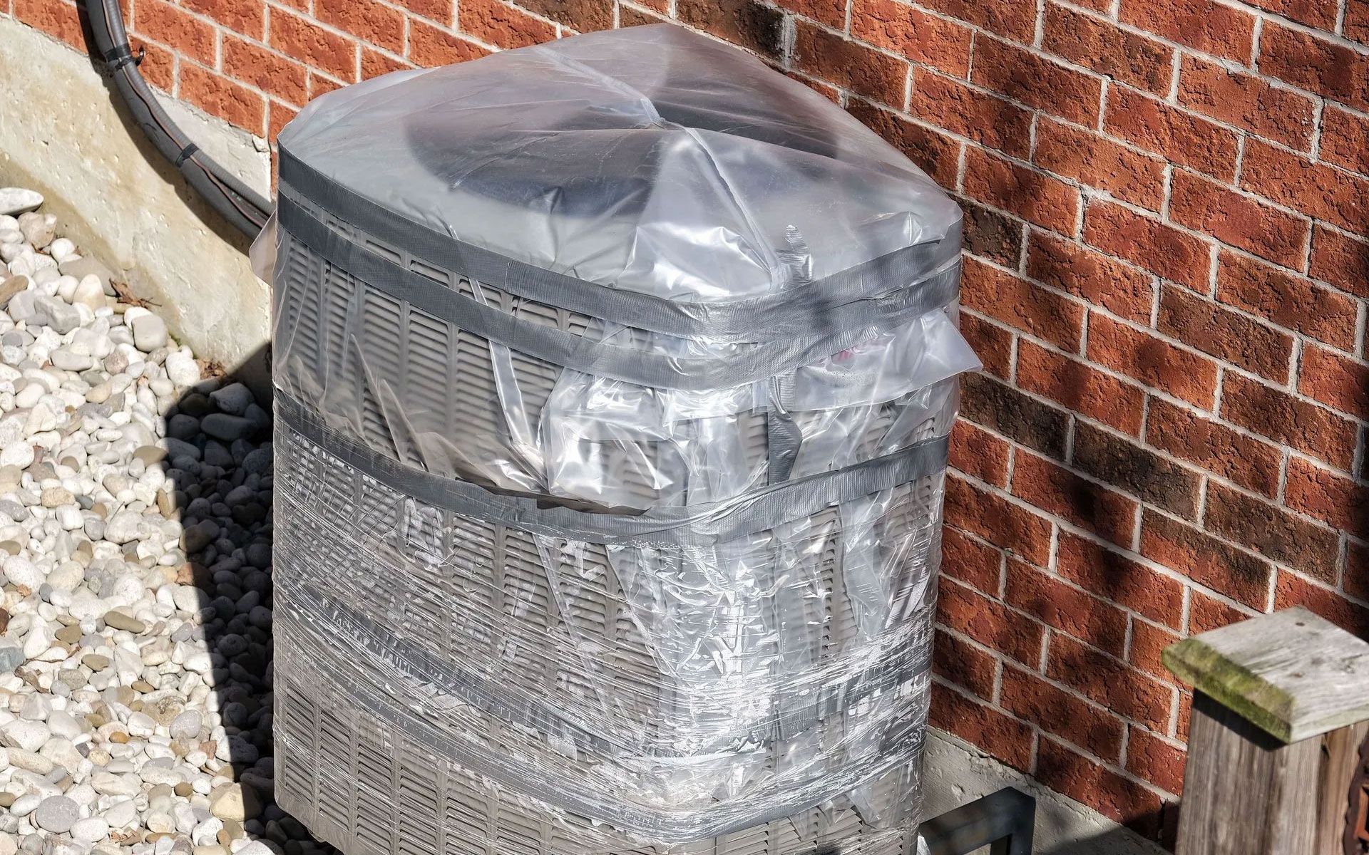 An outdoor air conditioning unit covered in plastic sheeting and secured with tape against a brick wall.