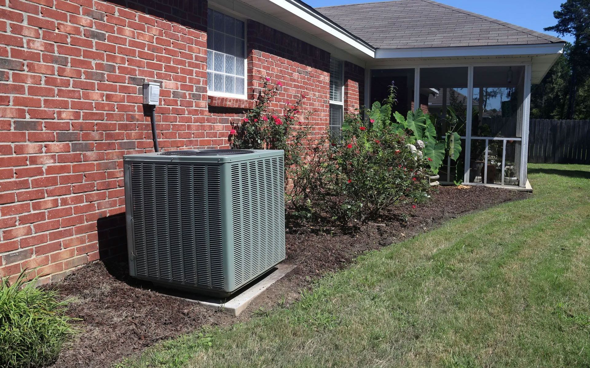 An outdoor air conditioning unit sits next to a brick house wall near a screened-in porch and garden beds.