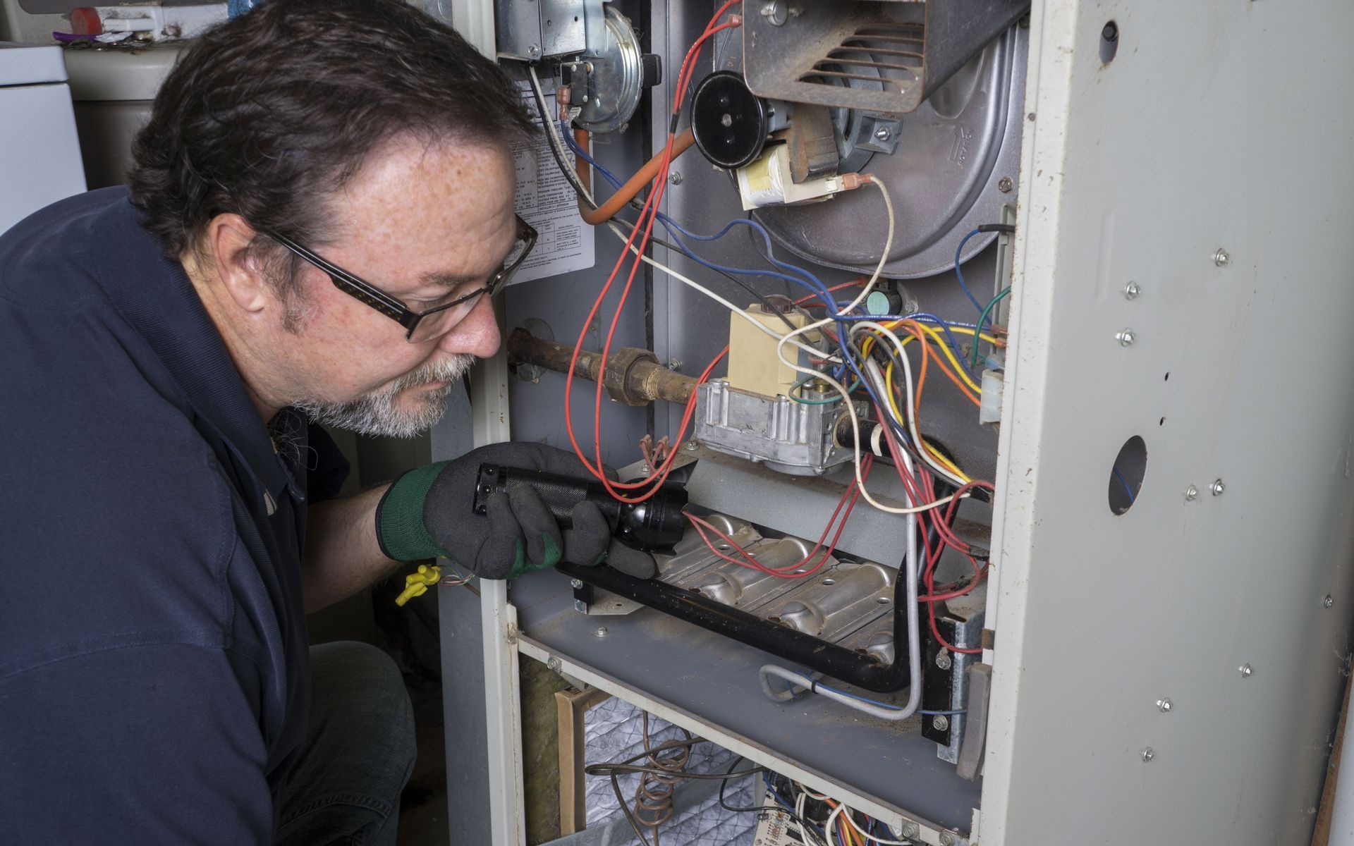A technician in a navy shirt and work gloves inspects the internal wiring and burner assembly of a home furnace.