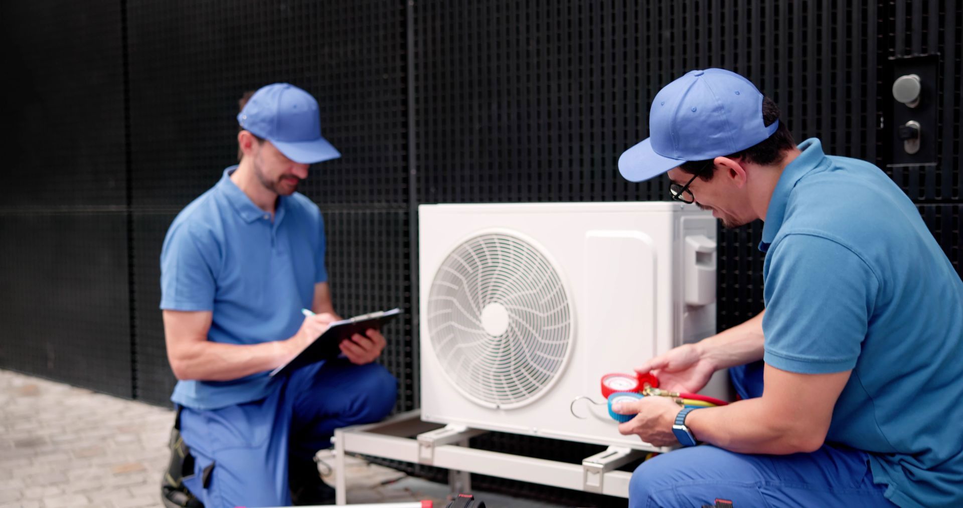 Two technicians in matching blue uniforms and caps service an outdoor air conditioning unit while one takes notes.