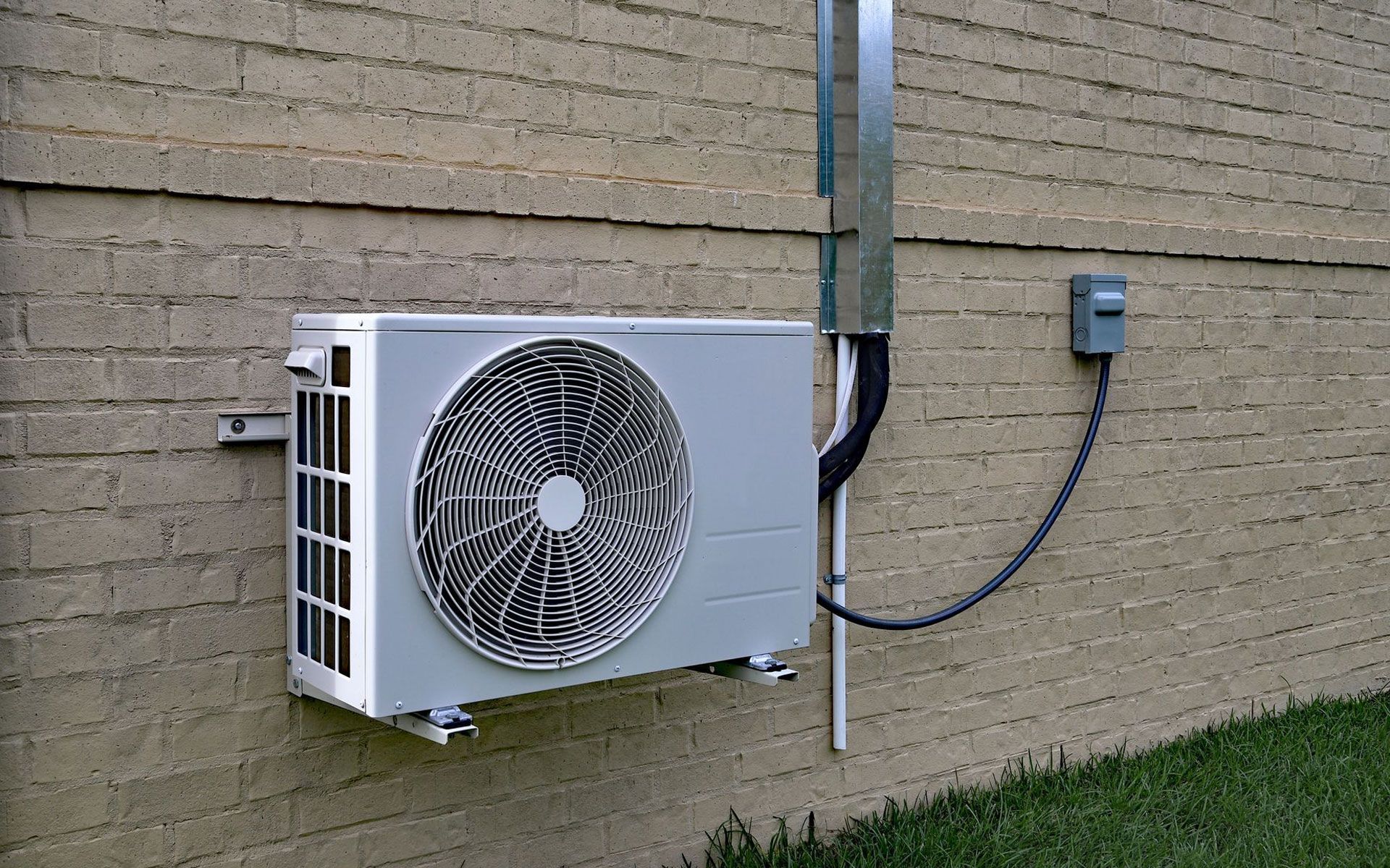 A white outdoor HVAC condenser unit mounted on a tan brick wall, connected to a nearby gray electrical disconnect box.