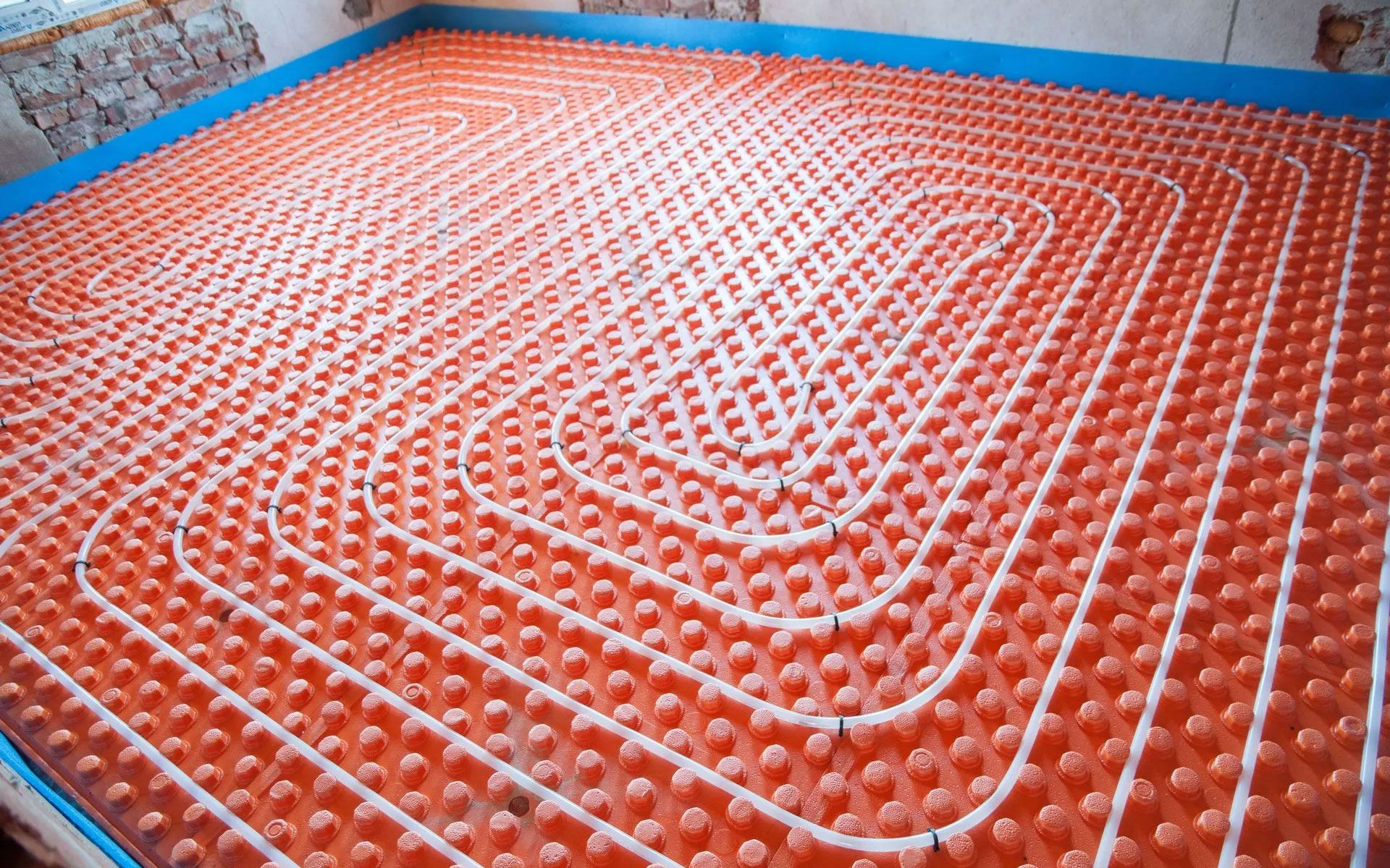 White radiant heating pipes laid in a spiral pattern on an orange dimpled insulation board across a room floor.