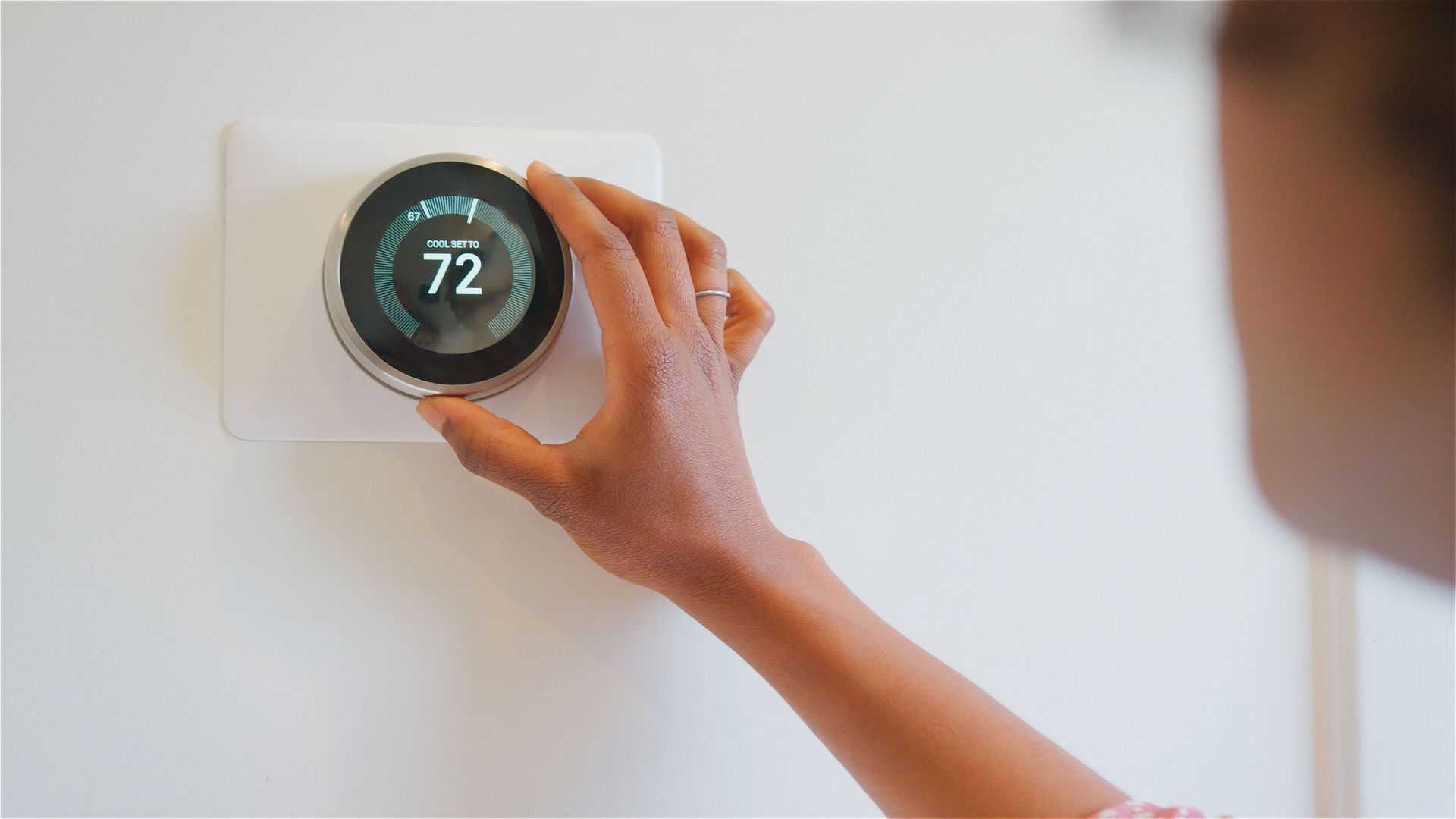 A hand adjusting a circular smart thermostat mounted on a white wall, displaying the temperature 72 degrees.