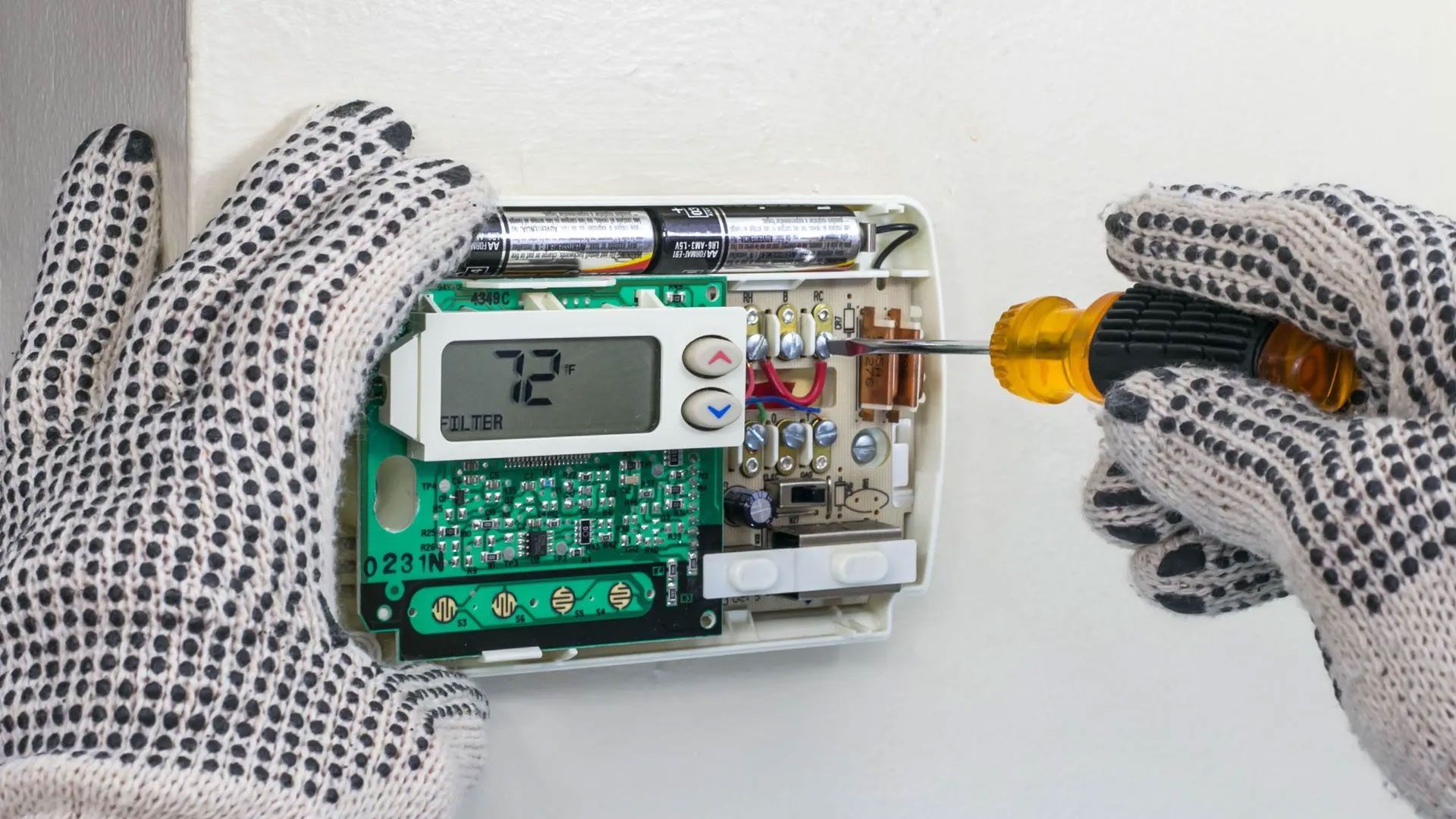 Gloved hands use a yellow-handled screwdriver to secure a wire on an open wall-mounted digital thermostat.