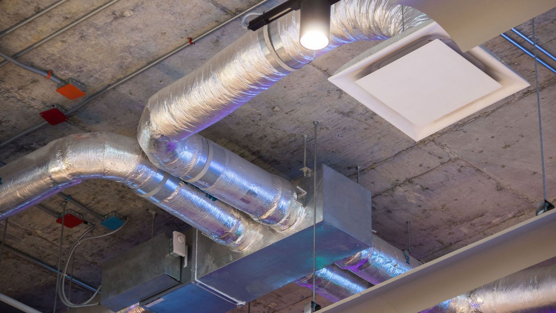 Exposed ceiling with silver ventilation ducts, a square white air diffuser, and conduit piping on a concrete surface.