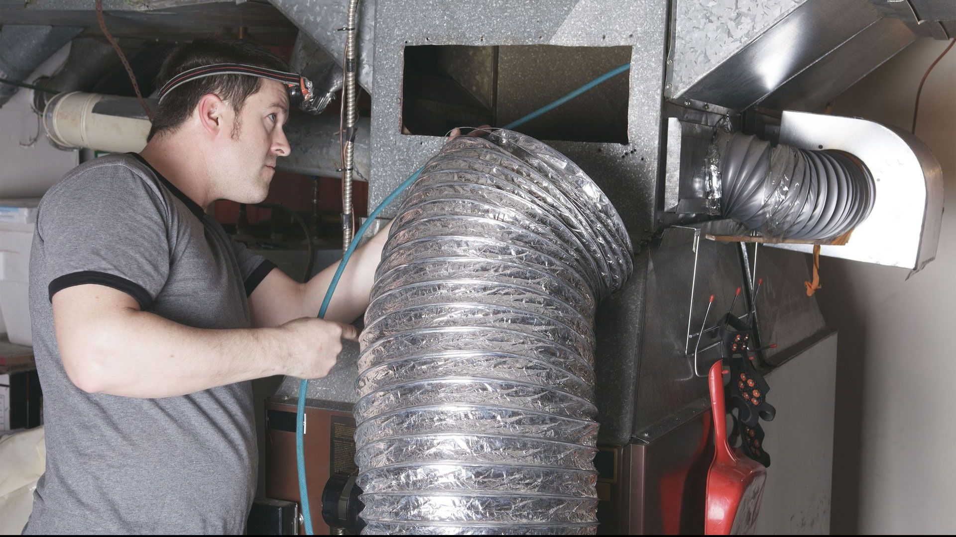 A person with a headlamp works on a silver flexible HVAC duct attached to a metal furnace in a basement.