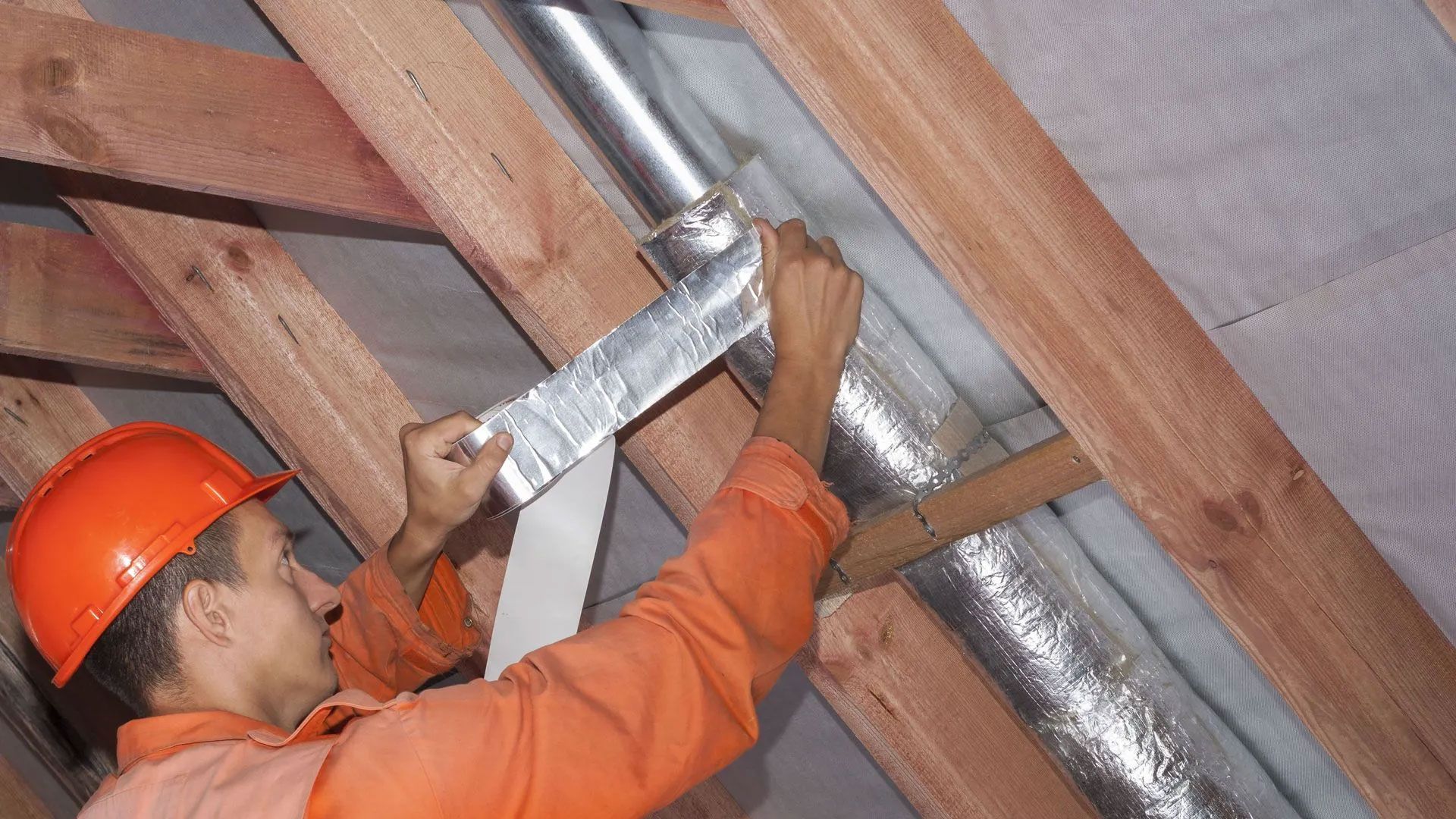 A worker in an orange uniform and hard hat applies silver tape to a metal ventilation duct in an unfinished attic.