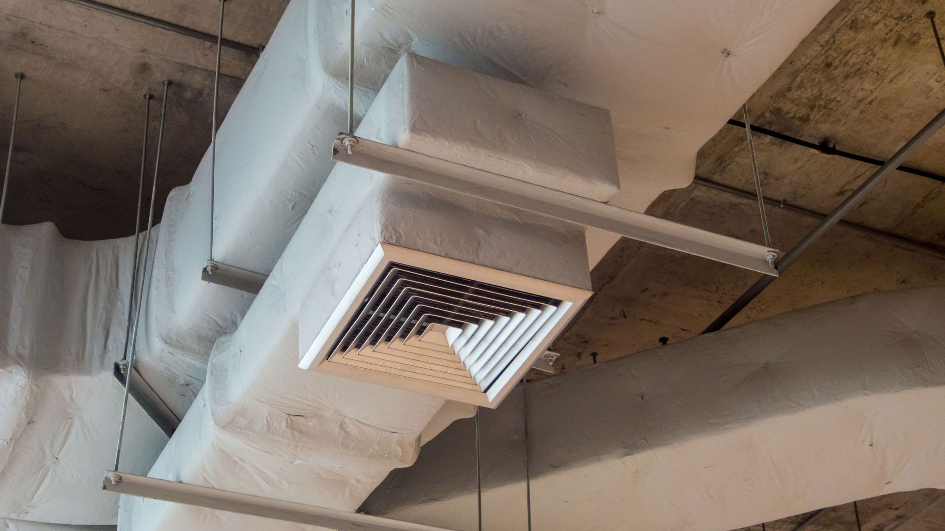 A square ventilation diffuser mounted on a white, insulated duct suspended from a concrete ceiling.