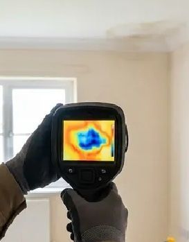 Gloved hands hold a thermal camera showing a heat map in a room with a window.
