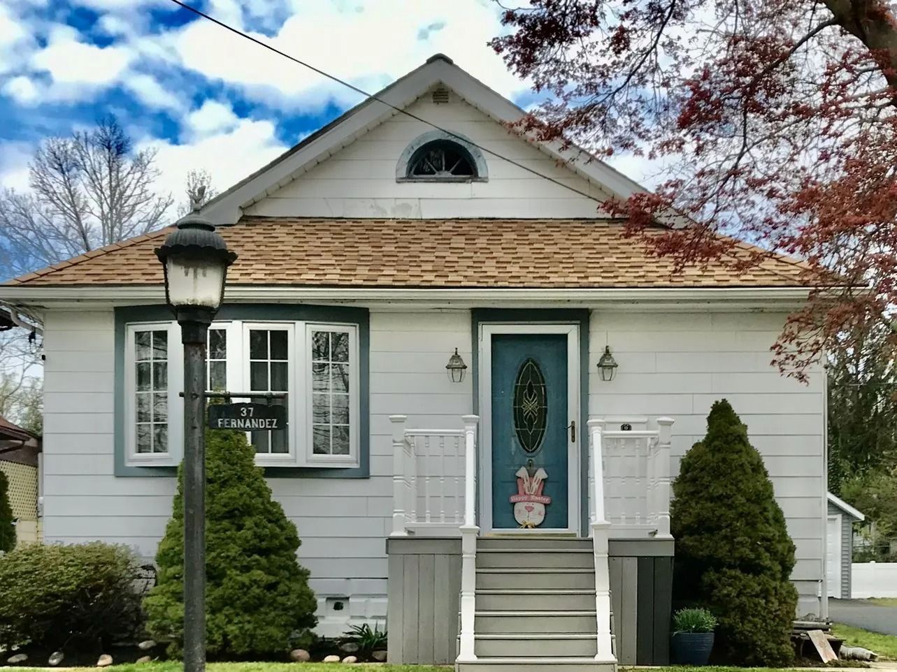 Small white house with teal front door, steps, shrubs, and a lamppost under a partly cloudy sky