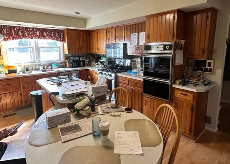 Cluttered kitchen with wooden cabinets, round table covered in papers, and sunlight from a window