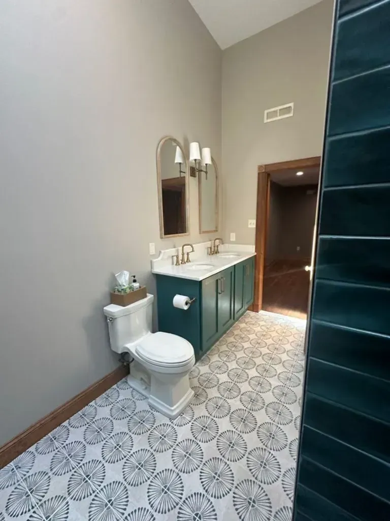 Small bathroom with patterned tile floor, teal vanity, toilet, mirror, and tall window above the doorway.