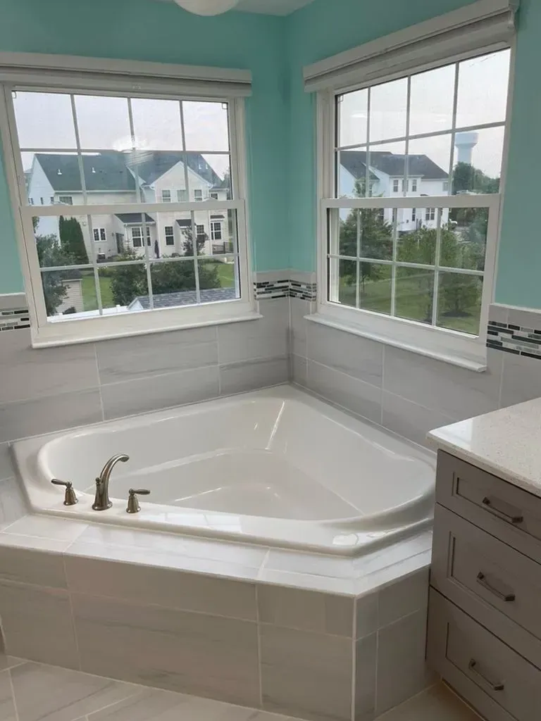 Bright bathroom with a white corner soaking tub, gray tile, and windows overlooking houses