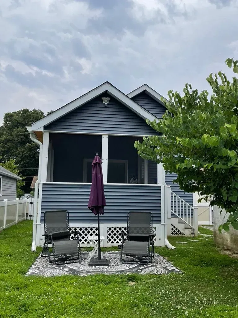 Small blue house with a front porch, two chairs, and a closed purple umbrella on a gravel patio.