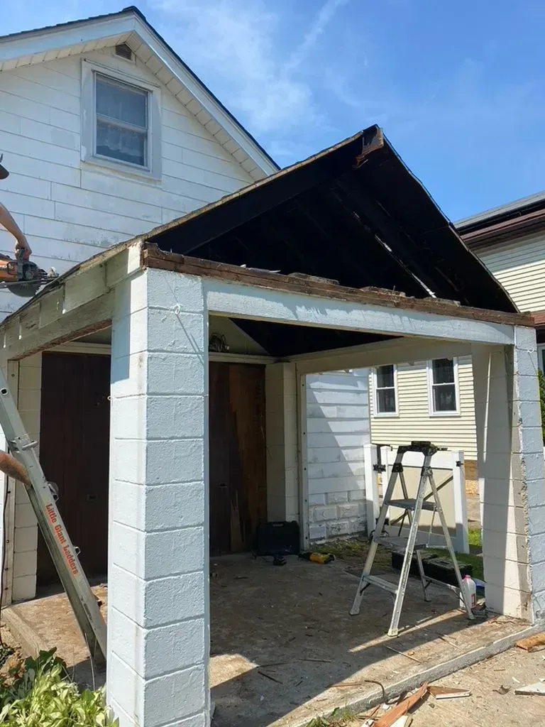 Partially built carport with white columns, dark roof, and a ladder beside a house under renovation
