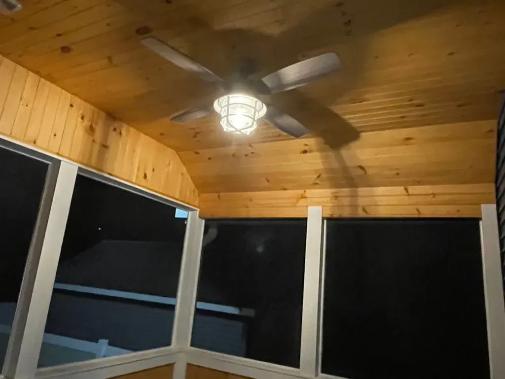 Wood-paneled screened porch ceiling with a lit fan, framed by dark window panels at night