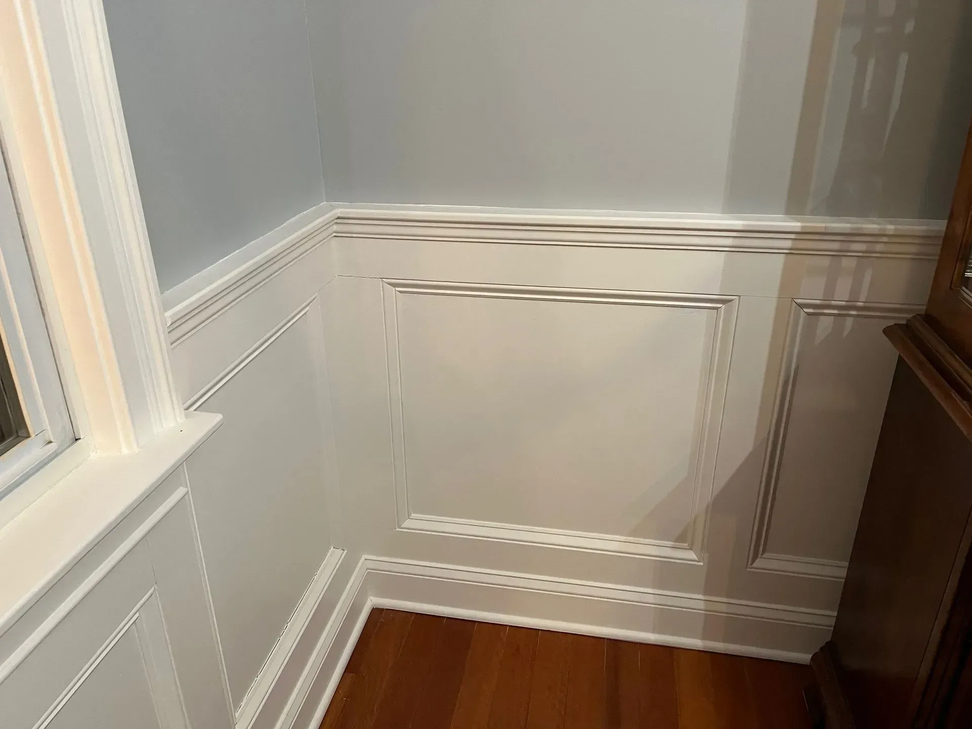 White paneled wall corner with molding and a hardwood floor beside a window and wooden cabinet