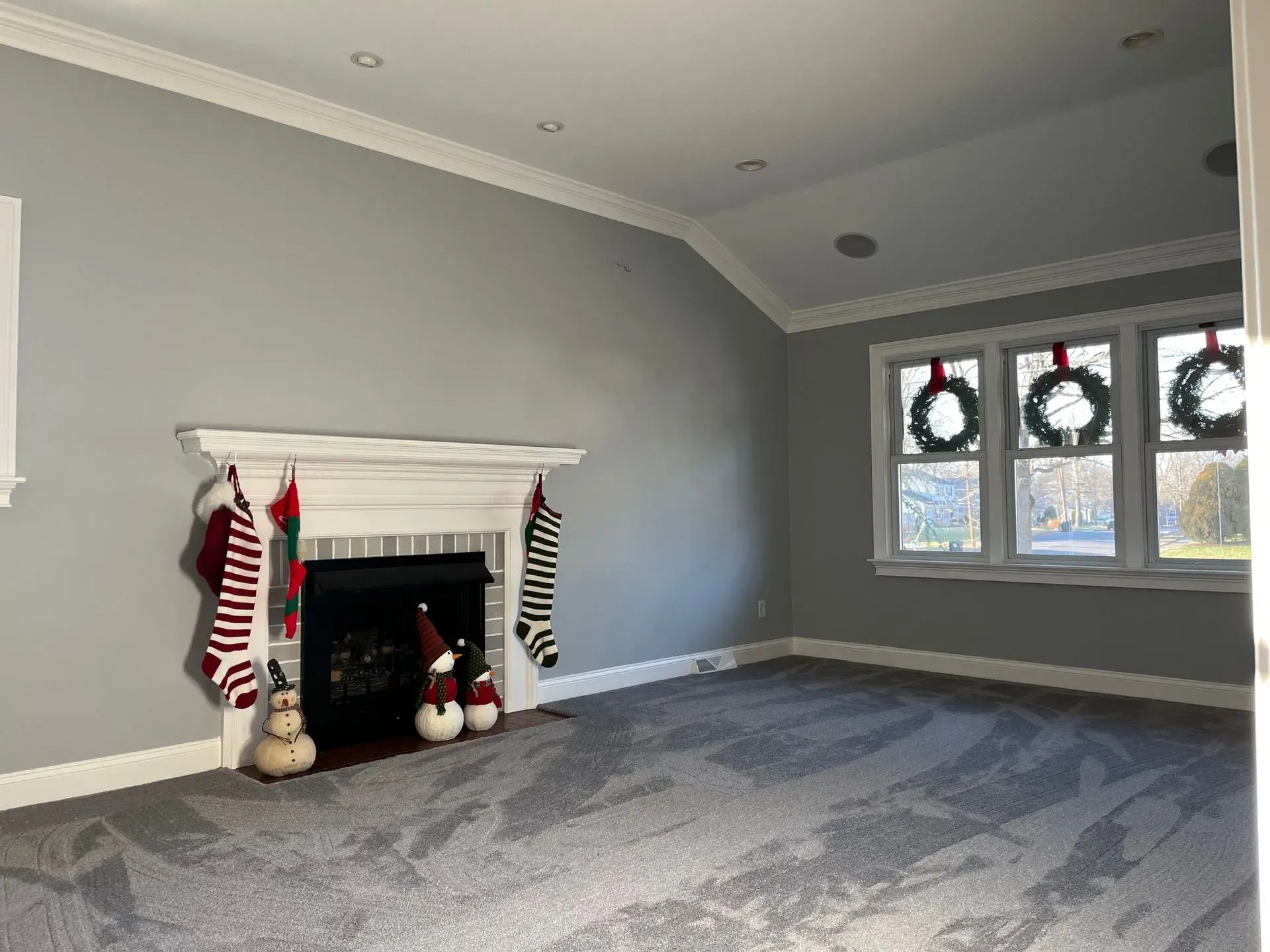 Empty living room with gray walls, fireplace, carpet, and three wreath-decorated windows.
