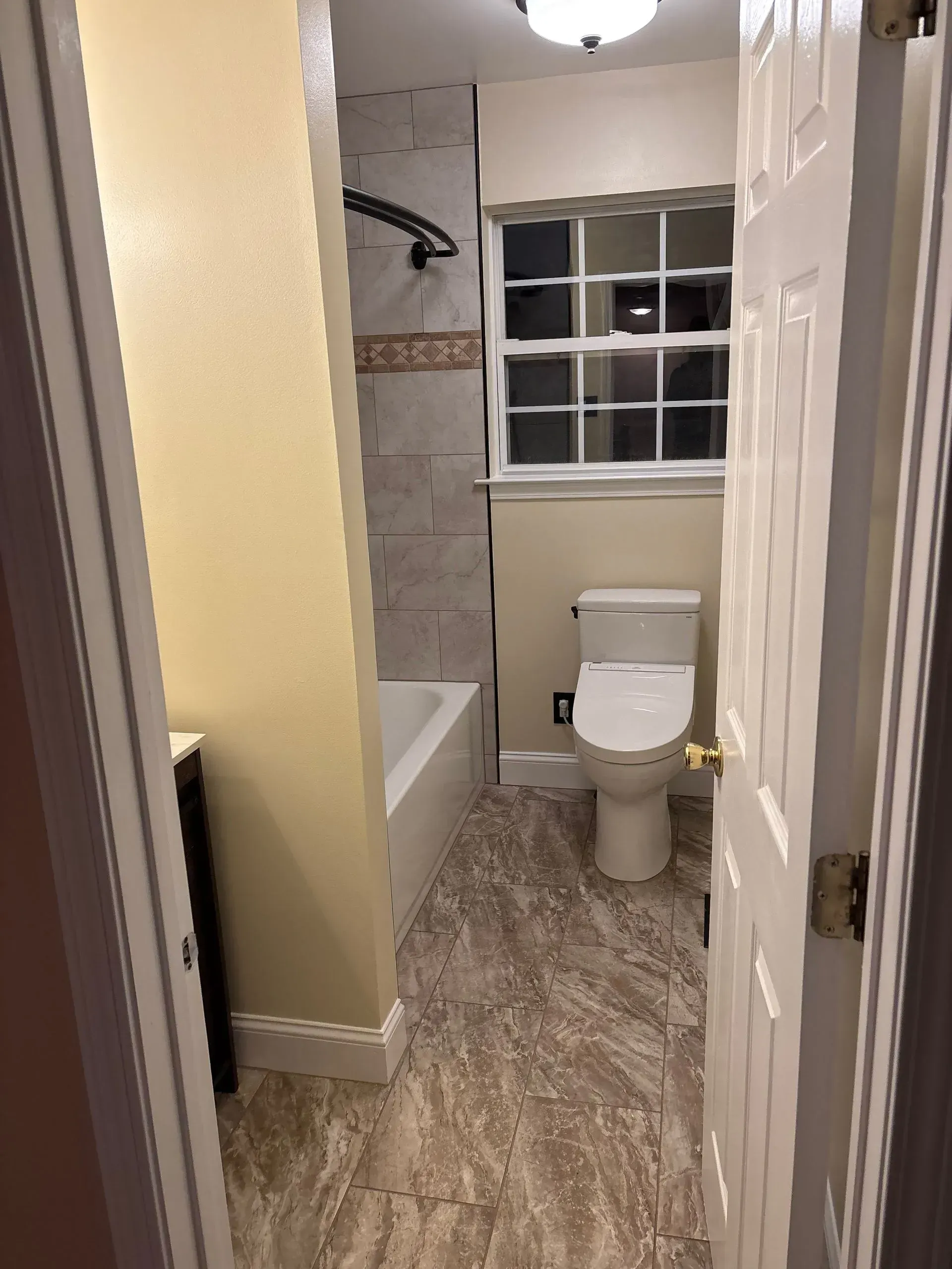 Narrow bathroom with toilet, bathtub, window, and beige walls, viewed from the doorway