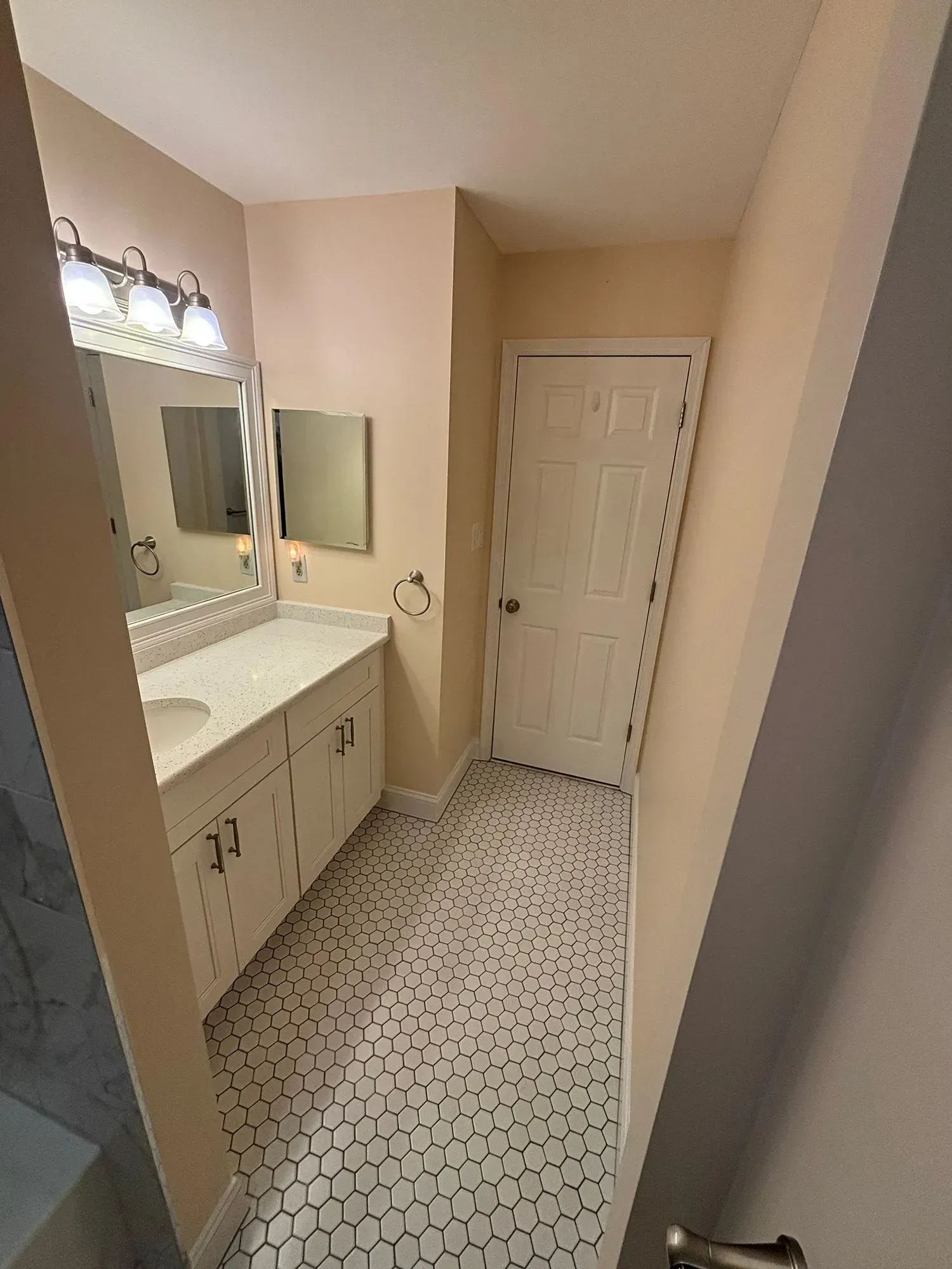 Narrow bathroom with white vanity, mirror, and patterned tile floor, viewed toward a closed door.