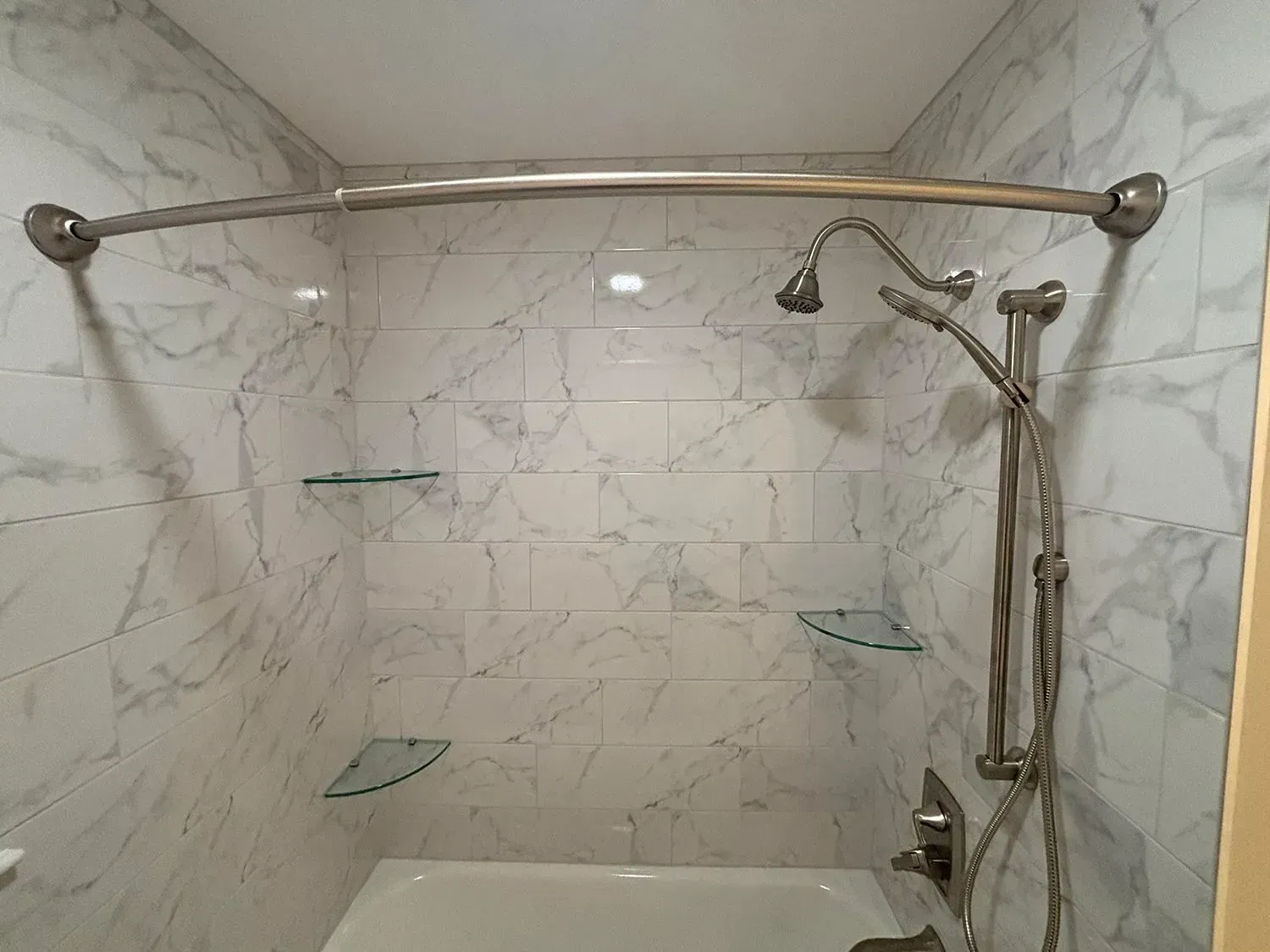 Walk-in shower with marble tile, curved curtain rod, glass shelves, and handheld showerhead