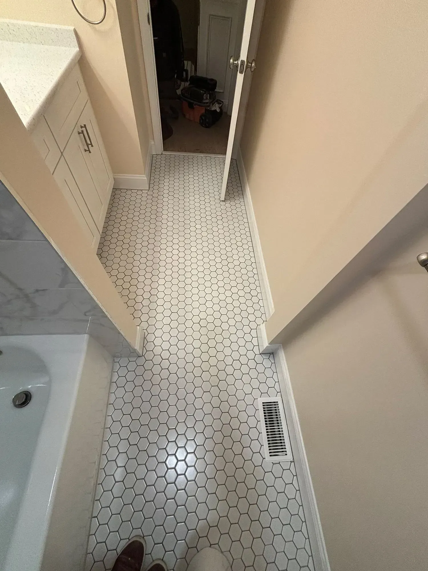 Narrow bathroom hallway with white hex tile floor, beige walls, sink at left, and open door ahead.