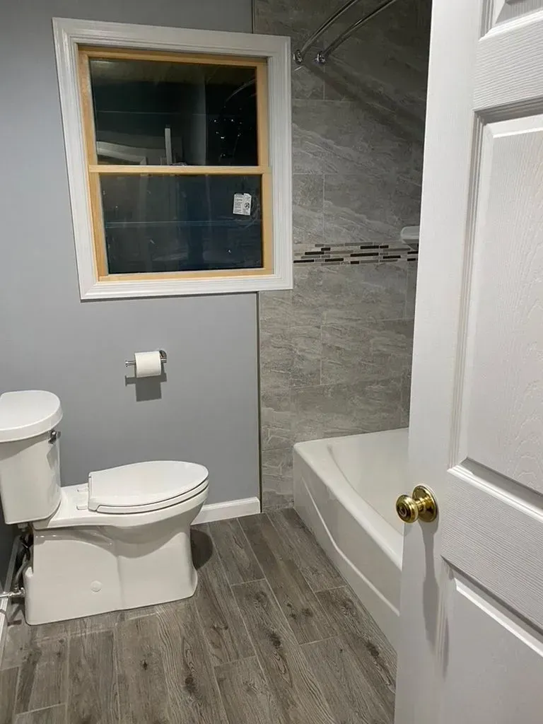 Small bathroom with toilet, window, gray walls, wood-look floor, and tiled shower stall.