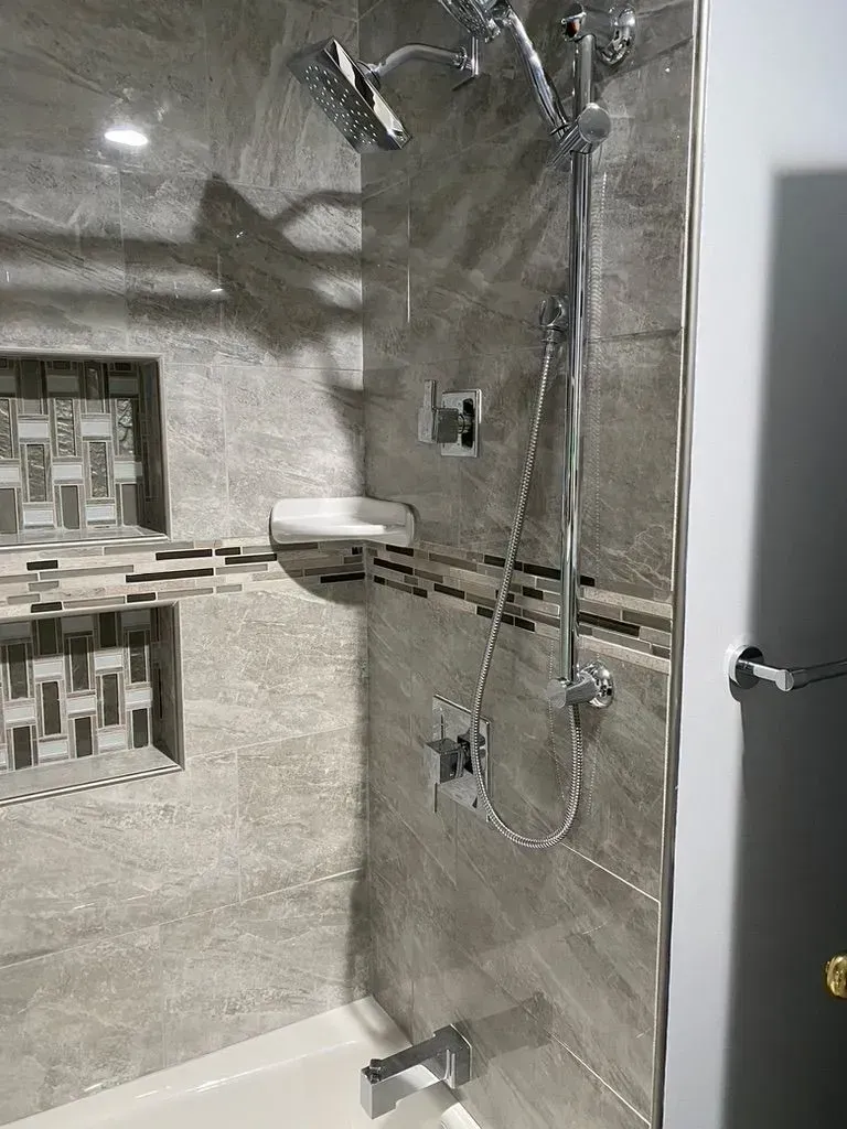 Bathroom shower with gray tiled walls, built-in shelf, and chrome showerhead/controls