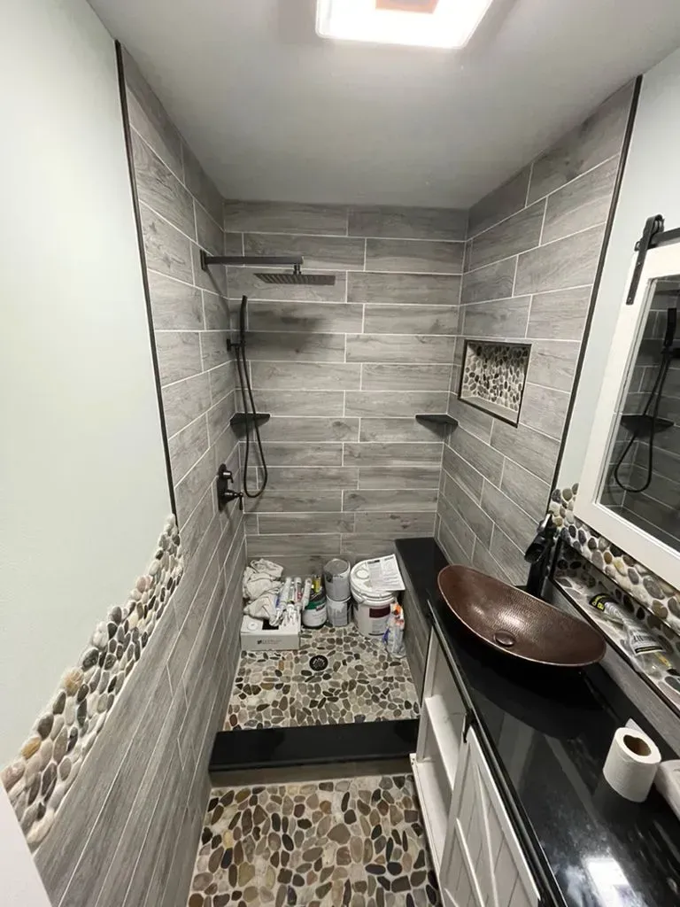 Narrow tiled bathroom with shower, wall shelf, mosaic floor, and window over a small sink counter