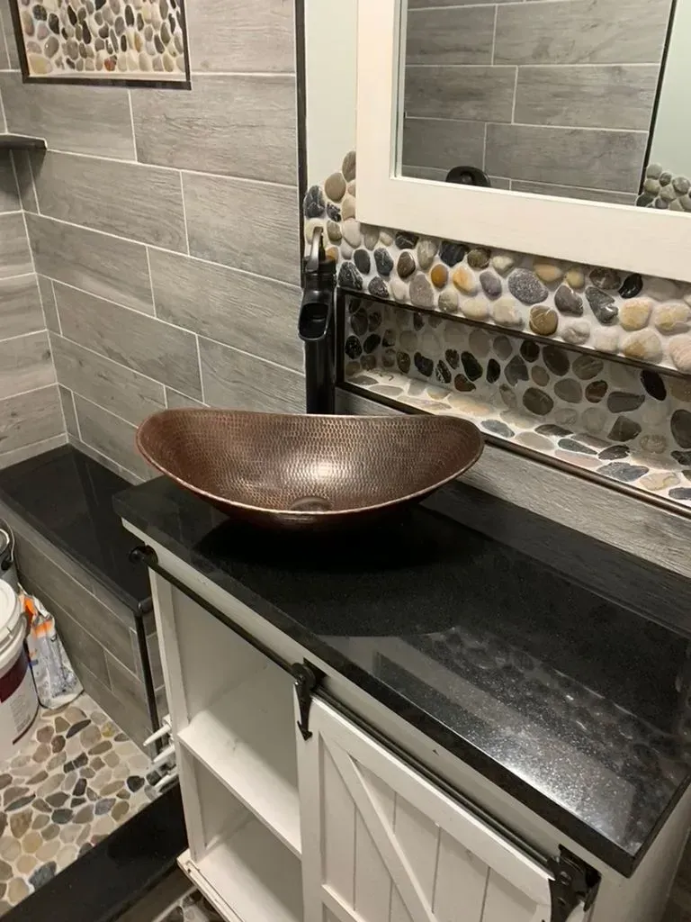 Bathroom vanity with black countertop, copper bowl sink, and gray tile walls