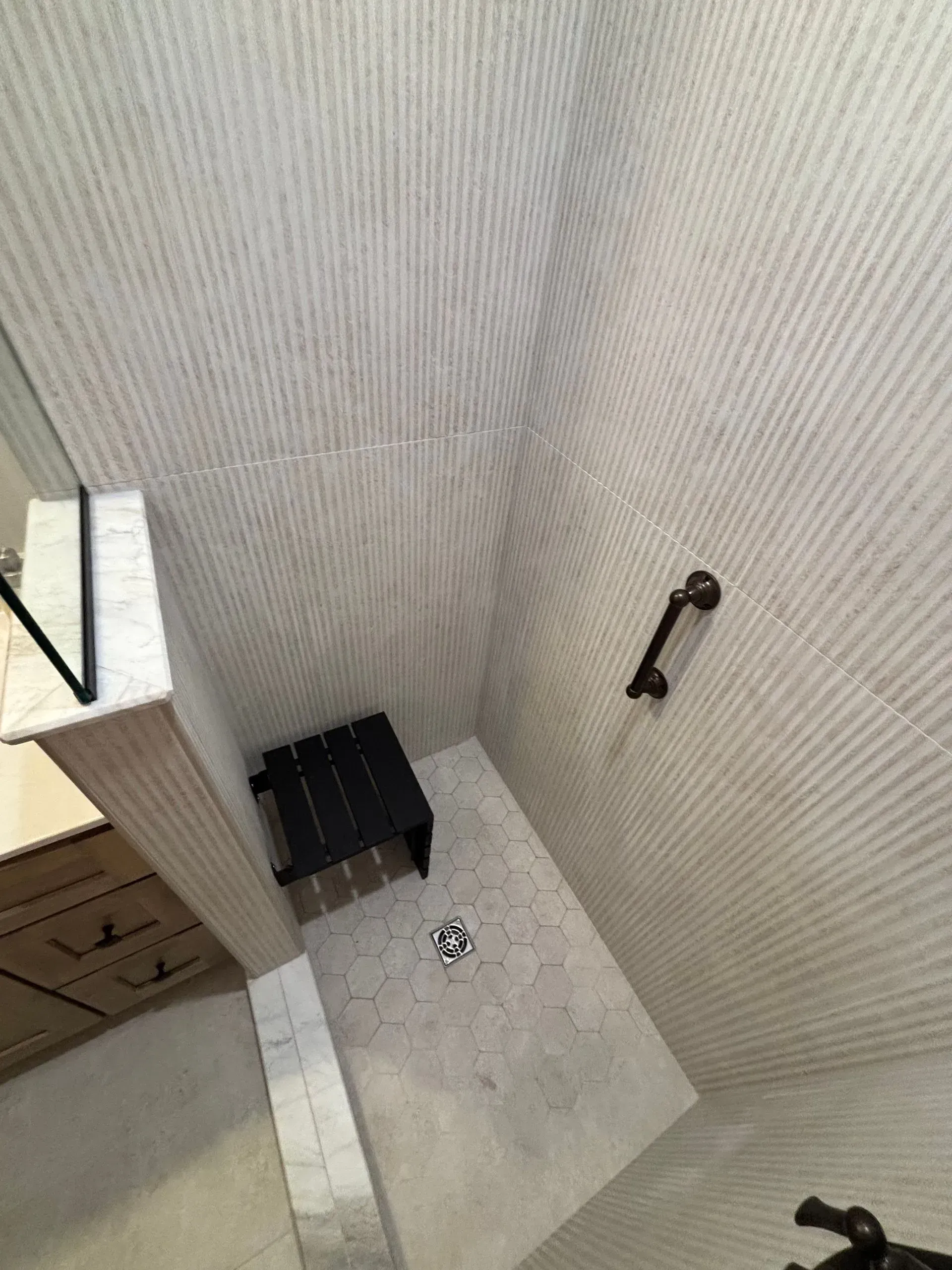 Small tiled shower corner with a black shelf, wall-mounted grab bar, and shower floor drain.