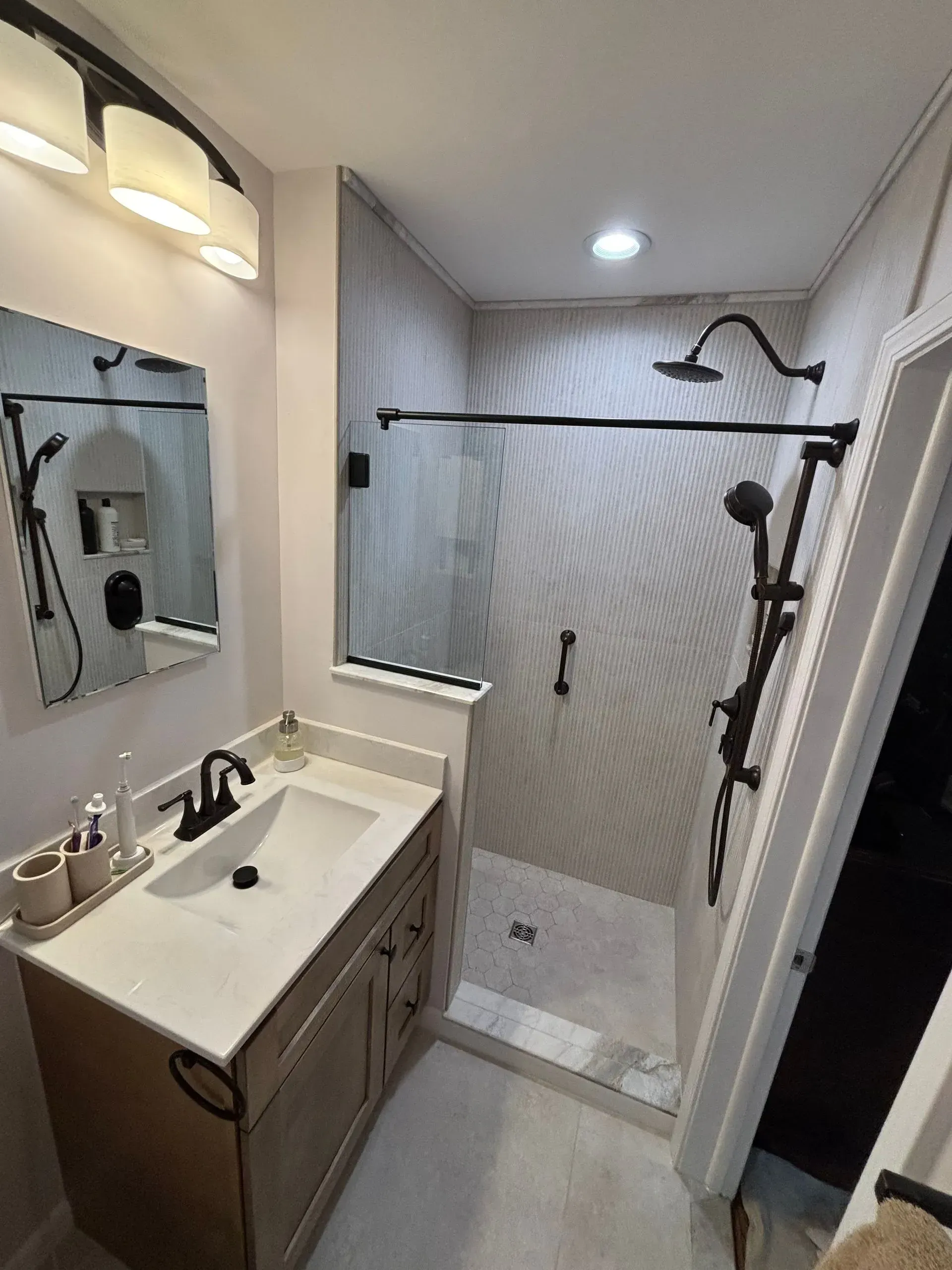 Small bathroom with vanity sink, mirror, shower bench, grab bars, and patterned shower curtain.