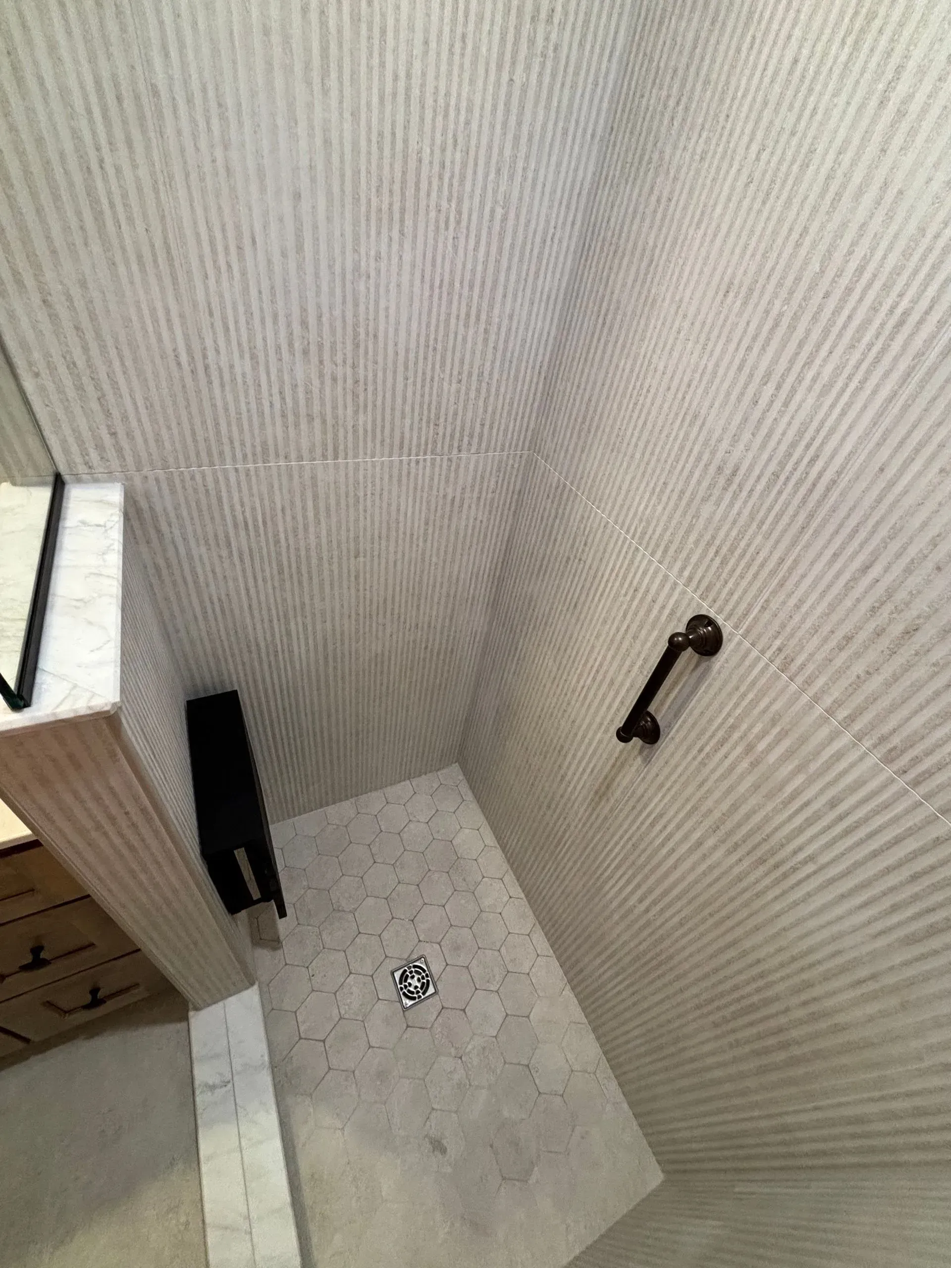 Small shower stall with beige tiled walls, white floor, drain, black shower head, and shelf on the left.