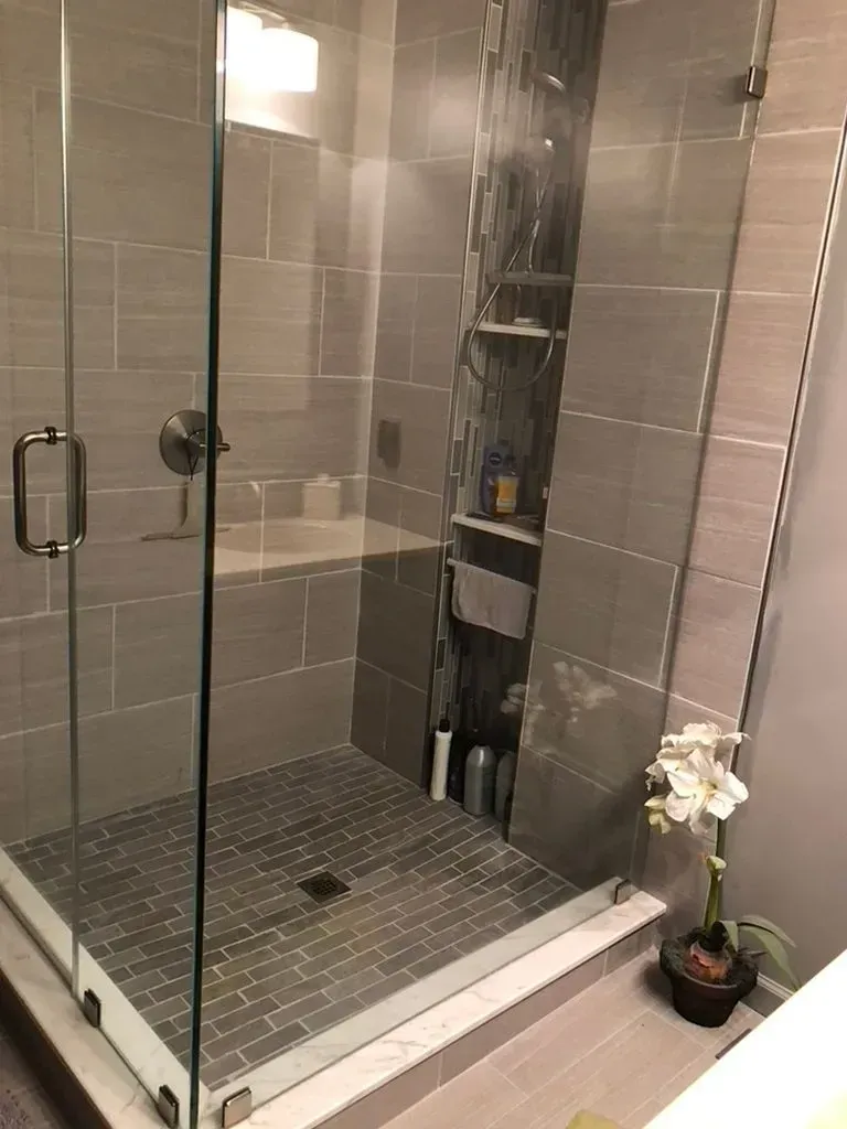 Glass walk-in shower with gray tile walls, pebble floor, and a potted orchid outside the door