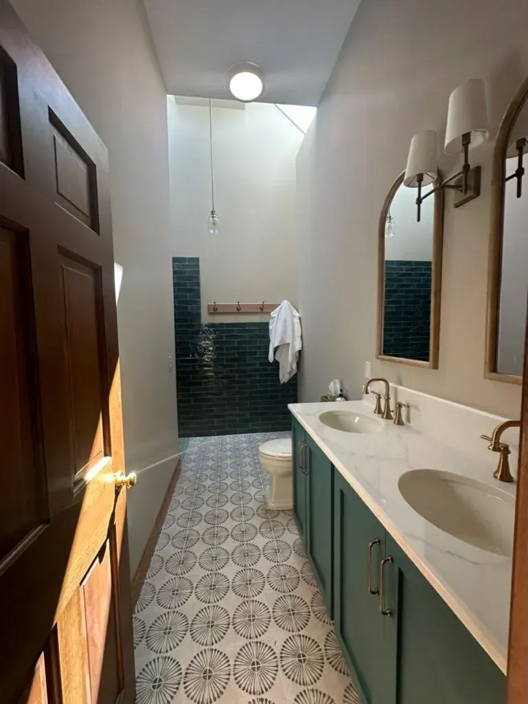 Narrow bathroom with blue tile, patterned floor, sink vanity, mirror, toilet, and wooden door.
