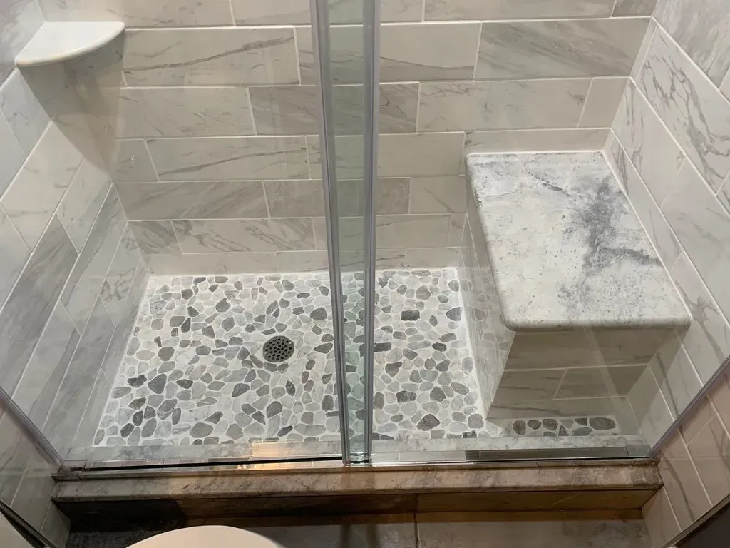 Glass shower with pebble floor and built-in corner bench, viewed through a partially open door.