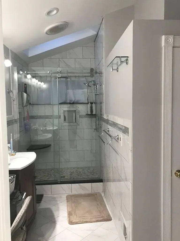 Small bathroom with glass shower, toilet, white tile walls, and a beige bath mat.