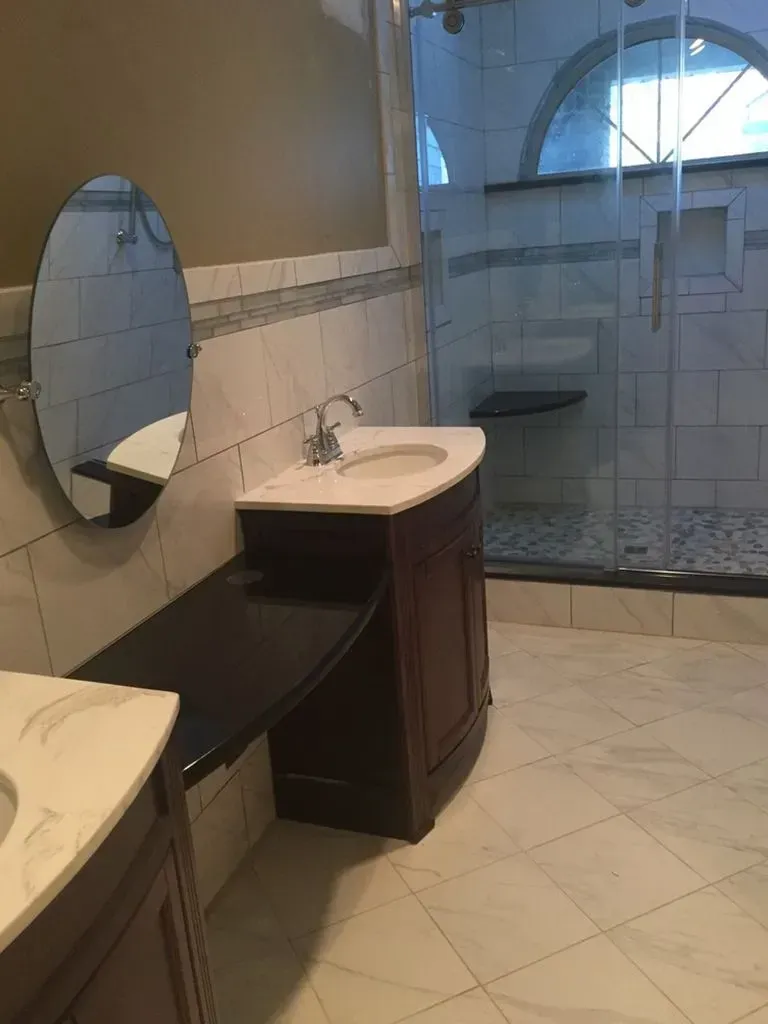 Bathroom vanity with oval mirror, sink, and tiled walk-in shower beyond glass door.
