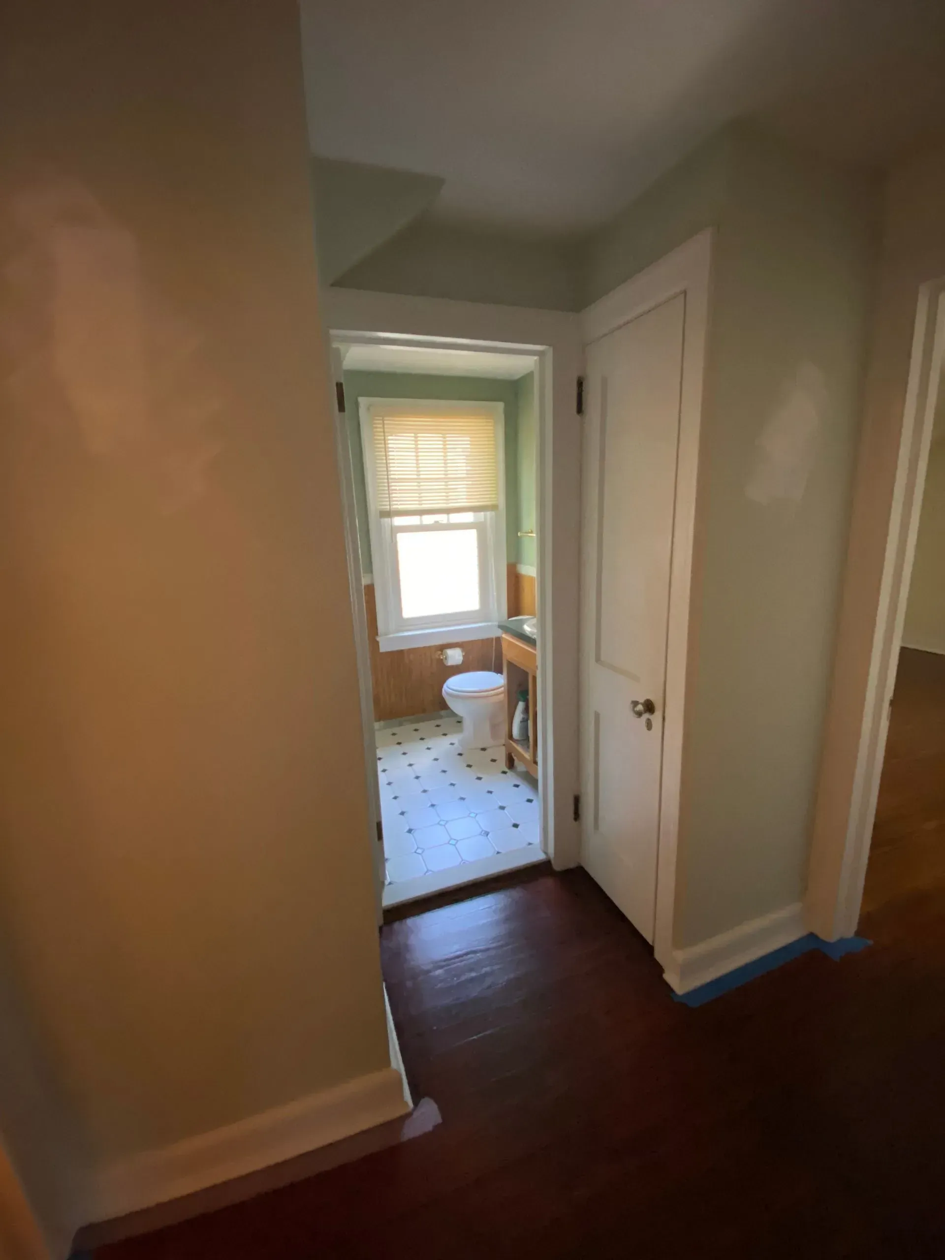 Narrow hallway with dark wood floors leading to a small tiled bathroom with a window.