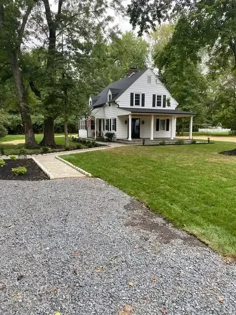 White house with a front porch and gravel driveway, surrounded by green lawn and trees