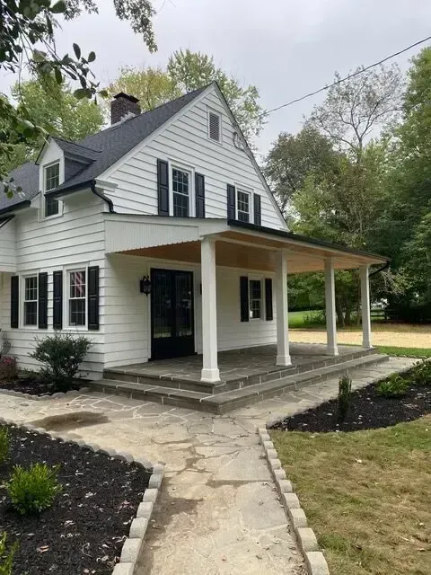 White two-story house with black shutters, front porch, and stone walkway surrounded by trees
